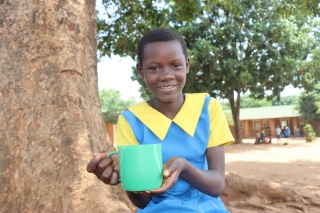 A girl, Ivy, stands next to a tree holding a mug of Mary's Meals and smiling