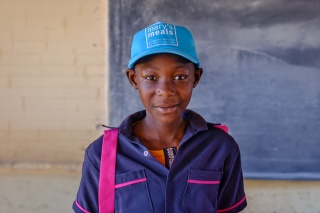 Tanaka, a young girl from Zambia smiling to camera with a dark blue and pink school shirt and a light blue Mary's Meals cap