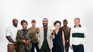 A group of celebrities stand against a white background. Stuart Douglas stands in front holding a coffee cup and a set of keys