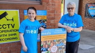 A woman and a girl stand in Mary's Meals t-shirts next to a Mary's Meals sign