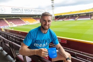 Stephen O'Donnell sits in a football stadium wearing a Mary's Meals t-shirt and holding a Mary's Meals mug