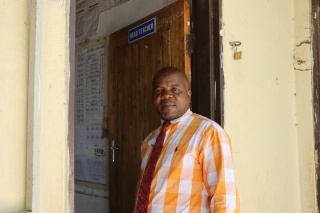 A man, Tiyese Tepete, stands outside a headteacher's office in a school