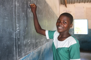 A child smiles whilst writing on a blackboard in the classroom