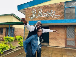 A young man with his arms out smiles while he piggy backs on a young woman's back, who laughs